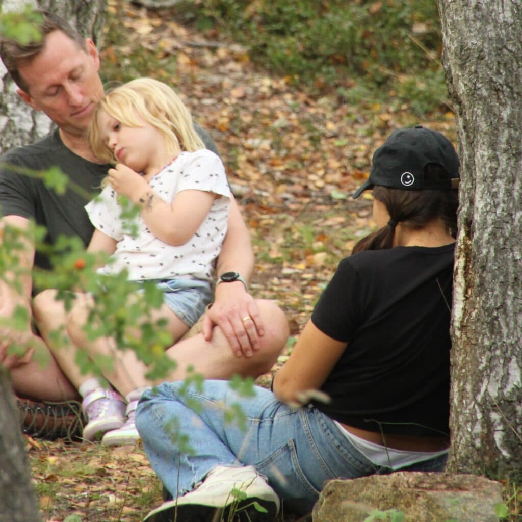 Bilden visar en grupp människor som sitter tillsammans utomhus i en skogsmiljö. Det ser ut som en ...